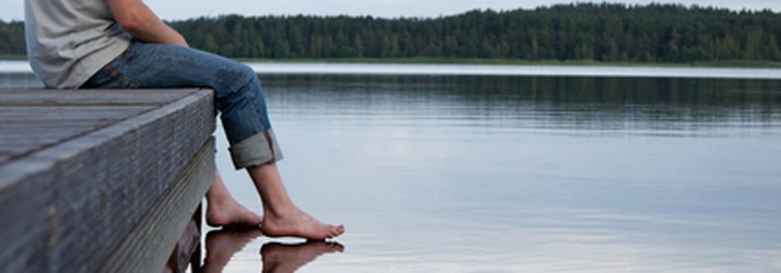 A person sits on a wooden dock with their feet in a calm lake, surrounded by a forested landscape, evoking a serene and tranquil atmosphere.