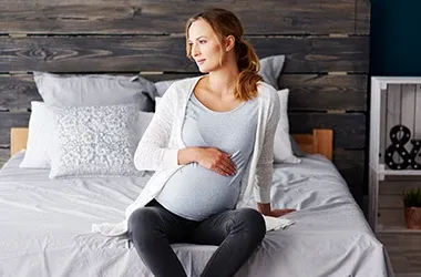 A pregnant individual is seated on a bed, gently holding their abdomen, in a cozy indoor setting.